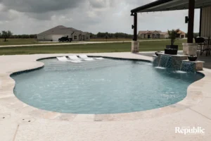 A modern, inviting pool with a waterfall feature, surrounded by a spacious patio and lush green lawn under a partly cloudy sky